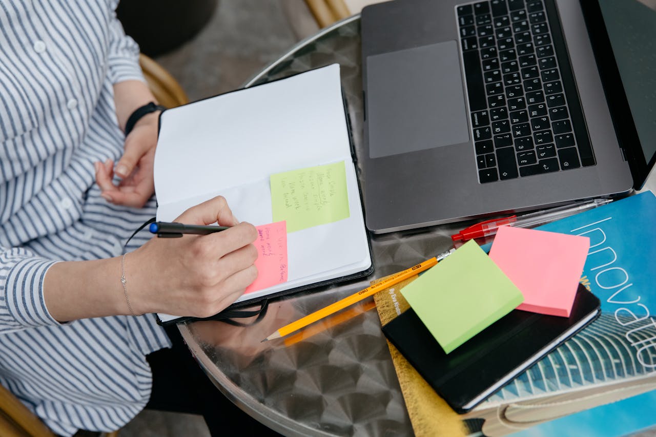 Person writing in notebook with laptop and sticky notes on a desk. Creative and organized workspace.