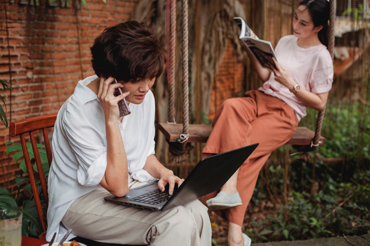 Pensive Asian female browsing laptop and talking on smartphone near resting Asian female reading magazine in garden near brick building in daytime