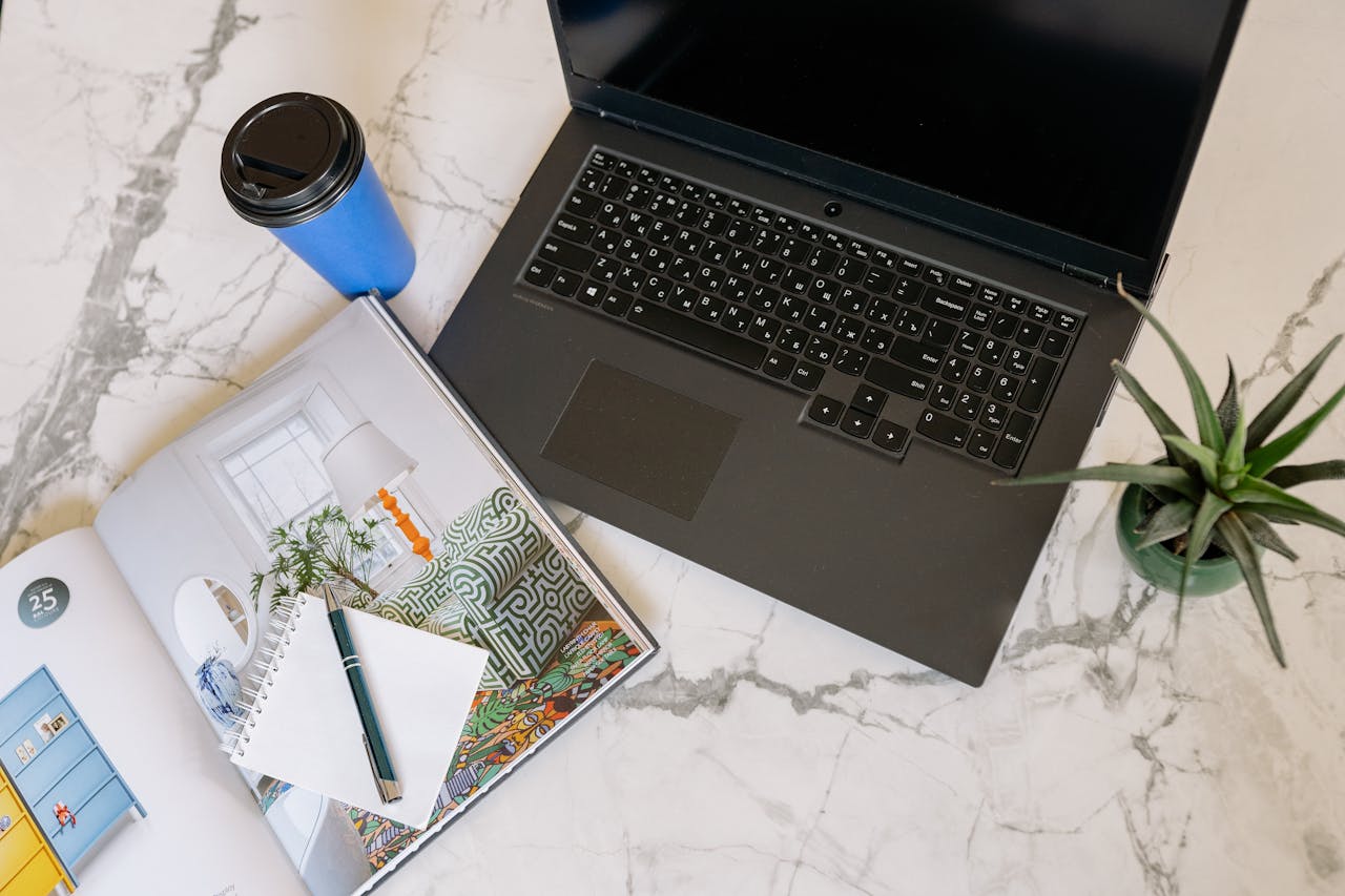 Overhead view of a modern workspace with a laptop, magazine, notepad, pen, and coffee cup on a marble desk.